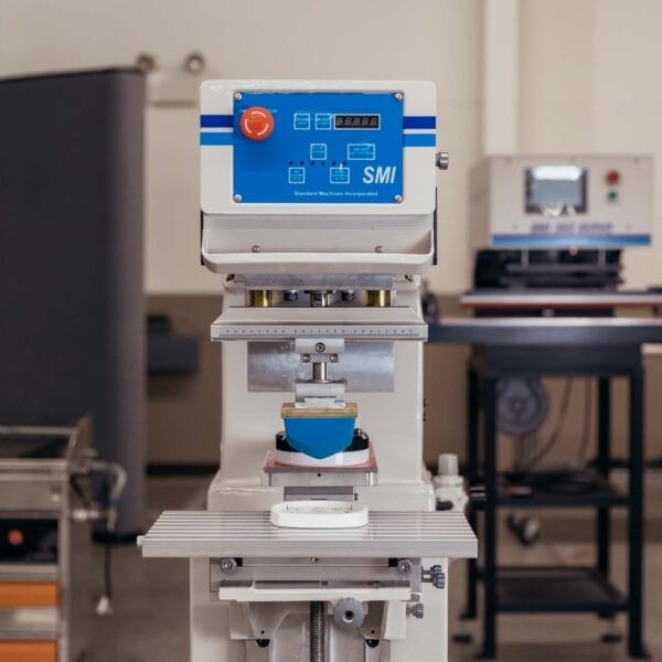 Front view of pad printing machine with blue silicone pad and digital control panel inside a manufacturing workshop.